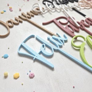 Könnte beinhalten: Eine Auswahl an Tortenaufsätzen in Schreibschrift, in Gold, Rosa, Blau und Grün. Pastellfarbene Streusel sind um die Aufsätze herum verstreut. Die Aufsätze sind in verschiedenen Schreibschriften gestaltet, was eine festliche Atmosphäre schafft.