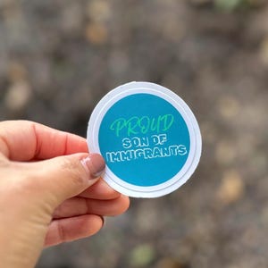 Può includere: Un adesivo rotondo con un bordo bianco e un centro blu. L'adesivo dice "Proud Son of Immigrants" in lettere verdi.