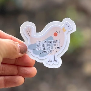 May include: A layered sticker in the shape of a bird, with a light pink body, blue wing, and orange beak. The sticker has text in Spanish: "¡POR FIN ENCONTRÉ AL PAJARITO QUE LE CONTABA TODO A MI MAMÁ!"