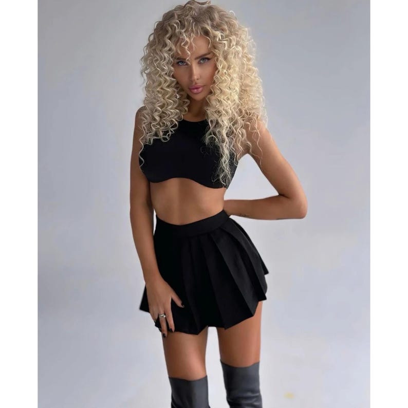 Op de afbeelding: Een vrouw draagt een zwart crop top en een zwarte geplooide rok. Ze heeft lang blond krullend haar en draagt zwarte laarzen.
