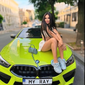 Op de afbeelding: Een vrouw zit op de motorkap van een felgroene sportwagen. De auto heeft een zwarte grille en een kentekenplaat met de tekst "MY RAY".