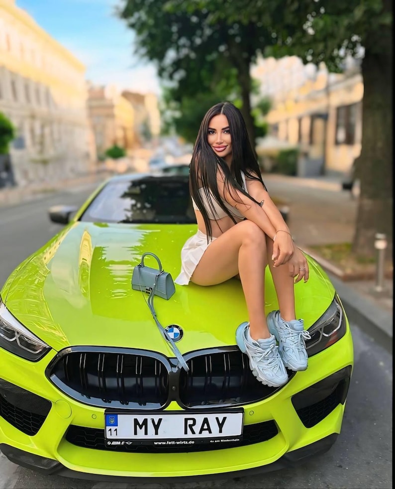 Op de afbeelding: Een vrouw zit op de motorkap van een felgroene sportwagen. De auto heeft een zwarte grille en een kentekenplaat met de tekst "MY RAY".