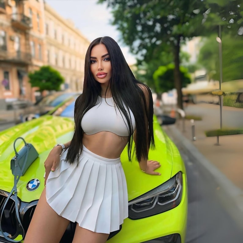 Op de afbeelding: Een vrouw in een wit topje en een witte geplooide rok, poseert naast een felgroene auto. Ze heeft lang zwart haar en kijkt naar de camera.