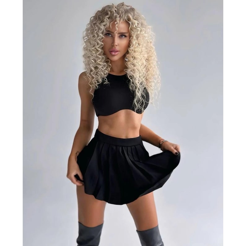 Op de afbeelding: Een vrouw draagt een zwart crop top en een zwarte geplooide rok. Ze heeft lang blond krullend haar en draagt zwarte laarzen.