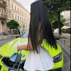 Op de afbeelding: Een vrouw in een witte geplooide rok staat naast een felgroene auto met een kentekenplaat waarop "MY RAY" staat.