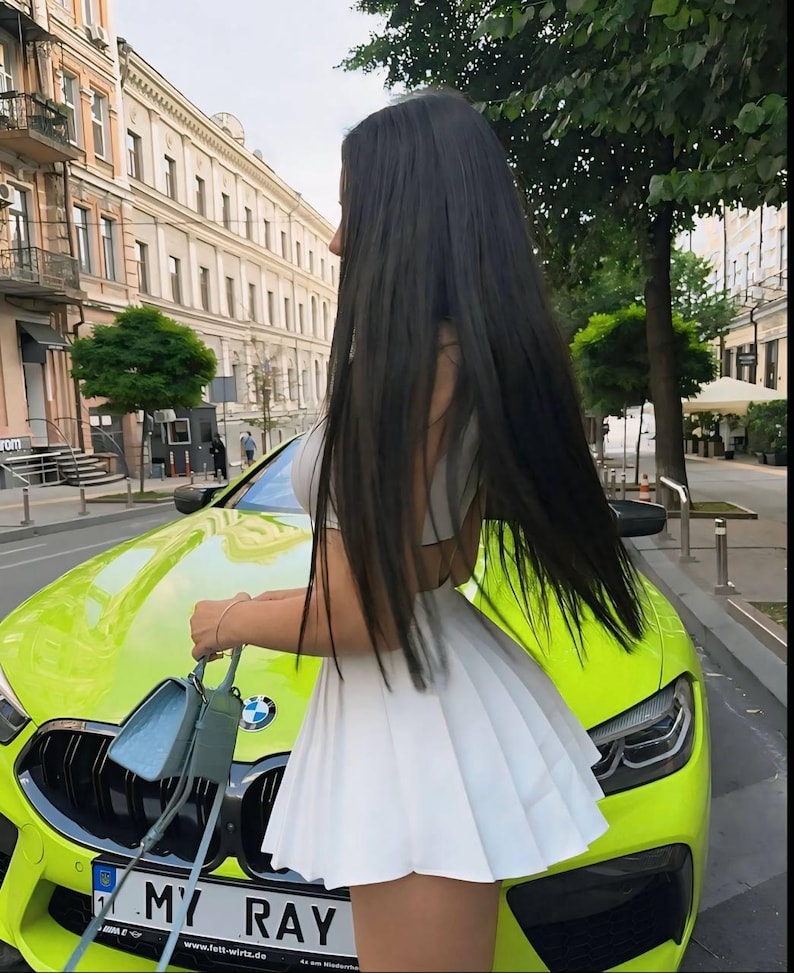 Op de afbeelding: Een vrouw in een witte geplooide rok staat naast een felgroene auto met een kentekenplaat waarop "MY RAY" staat.