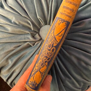 Pu&ograve; includere: Un libro d'epoca intitolato "Blithedale Romance" di Hawthorne. Il dorso del libro &egrave; di colore arancione chiaro con motivi decorativi blu e oro. Il libro &egrave; tenuto contro un cuscino di velluto blu chiaro.