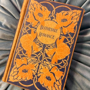 Pu&ograve; includere: Un libro vintage intitolato "Blithedale Romance" con copertina blu navy e design floreale arancione. Il libro &egrave; appoggiato su una superficie di velluto blu chiaro, creando un contrasto di colori e trame. Il titolo del libro &egrave; in una cornice circolare.