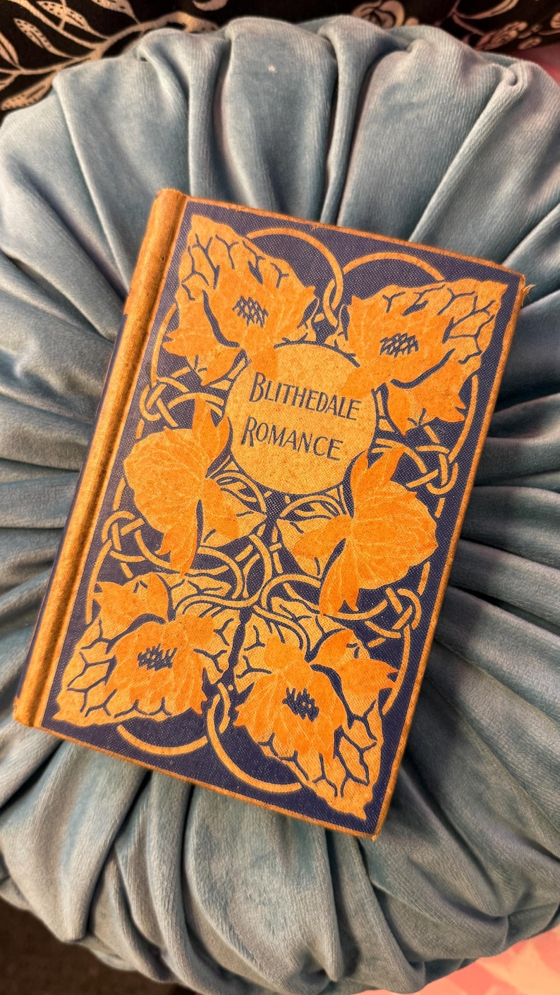 Pu&ograve; includere: Un libro vintage intitolato "Blithedale Romance" con copertina blu navy e design floreale arancione. Il libro &egrave; appoggiato su una superficie di velluto blu chiaro, creando un contrasto di colori e trame. Il titolo del libro &egrave; in una cornice circolare.