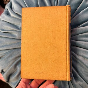 Pu&ograve; includere: Un libro vintage con una copertina color senape e un dorso blu. Il libro &egrave; tenuto in mano e poggia su un cuscino di velluto blu chiaro. La copertina ha un aspetto strutturato, che suggerisce et&agrave; e utilizzo.