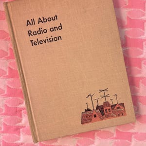 Könnte beinhalten: Beiges Hardcover-Buch mit dem Titel "All About Radio and Television". Das Buch zeigt eine kleine Illustration von Häusern mit Antennen. Es liegt auf einer rosa-weiß gemusterten Oberfläche.