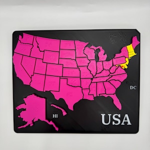 May include: A black rectangular map of the USA with each state outlined in bright pink. The states of Maine, New Hampshire, and Vermont are highlighted in yellow. The word "USA" is printed in white at the bottom. Braille dots are visible on the map.