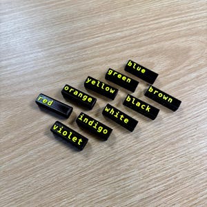 May include: Black rectangular tiles with the names of colours written in yellow letters. The colours are red, orange, yellow, green, blue, brown, black, white, indigo, and violet.