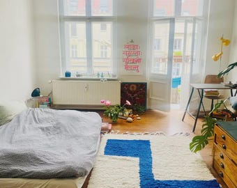 Puede incluir: Un dormitorio luminoso y espacioso con una cama cubierta con un edredón a cuadros grises y blancos. Una alfombra blanca con un llamativo diseño geométrico azul está sobre el suelo de madera. La habitación tiene grandes ventanas y un póster con texto.