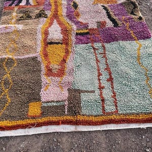 May include: A colorful, rectangular rug with a textured surface. The rug features a patchwork design with shades of brown, pink, purple, yellow, and green. The edges are trimmed with a red and orange border. The rug is likely made of wool or a similar material.