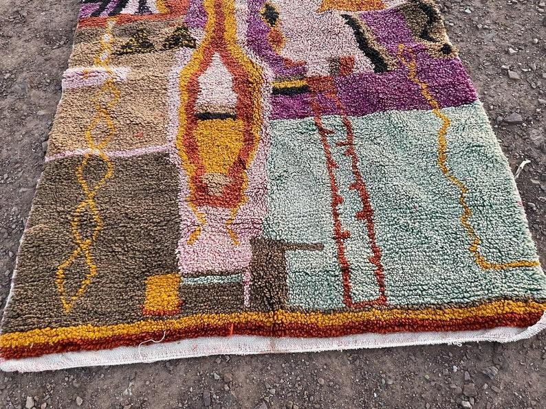 May include: A colorful, rectangular rug with a textured surface. The rug features a patchwork design with shades of brown, pink, purple, yellow, and green. The edges are trimmed with a red and orange border. The rug is likely made of wool or a similar material.