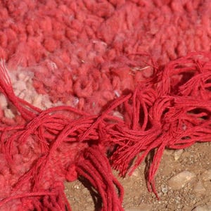 May include: Close-up of a vibrant red rug with a textured, woven surface. The rug features a mix of red and off-white hues, with long, knotted red fringe extending from the edge. The rug's design is detailed and intricate.