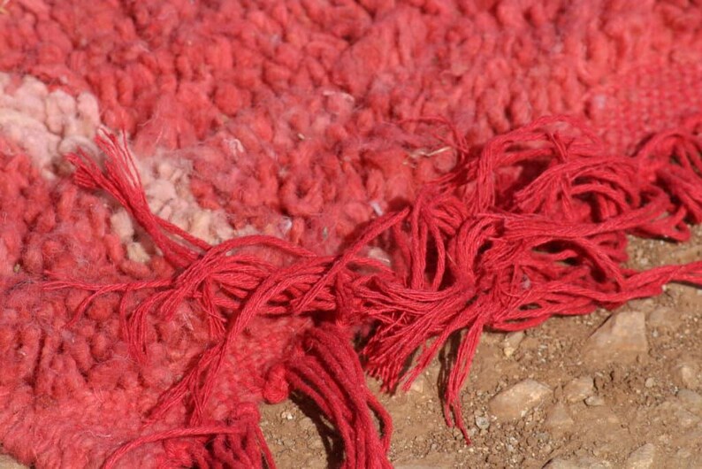 May include: Close-up of a vibrant red rug with a textured, woven surface. The rug features a mix of red and off-white hues, with long, knotted red fringe extending from the edge. The rug's design is detailed and intricate.