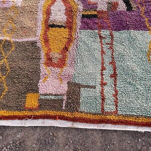 May include: A colorful, textured rug with a geometric design. The rug features a mix of brown, pink, yellow, purple, and green hues. The design includes rectangular and linear shapes, with a white border along the bottom edge.