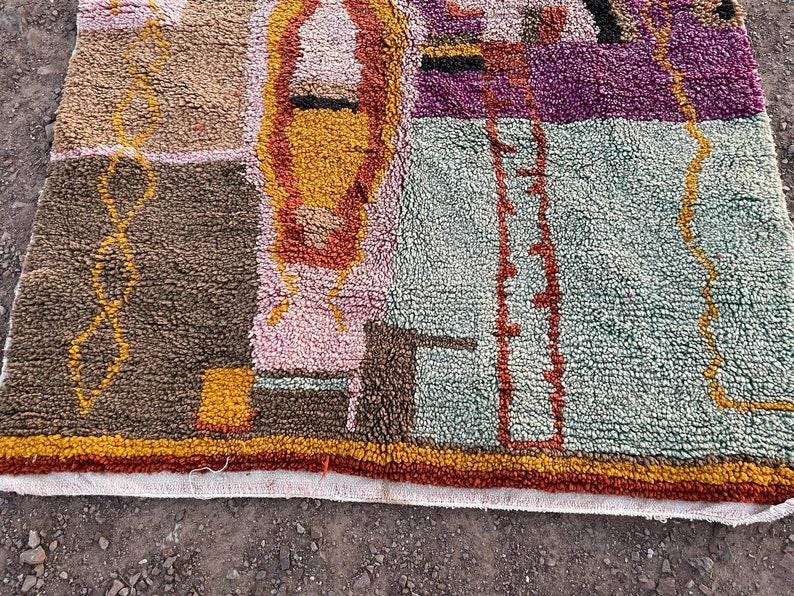 May include: A colorful, textured rug with a geometric design. The rug features a mix of brown, pink, yellow, purple, and green hues. The design includes rectangular and linear shapes, with a white border along the bottom edge.