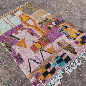 May include: A colorful, rectangular rug with a geometric design. The rug features a mix of colors, including pink, purple, yellow, and black. It has a fringe along one edge and is made of a textured material.