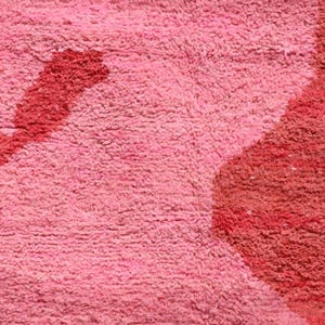 May include: Close-up of a pink and red textured rug. The rug has a soft, plush appearance with a pattern of red shapes and lines against a pink background. The texture is dense and appears to be made of a looped material.
