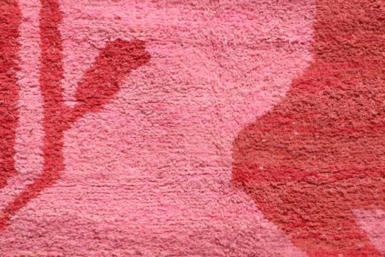 May include: Close-up of a pink and red textured rug. The rug has a soft, plush appearance with a pattern of red shapes and lines against a pink background. The texture is dense and appears to be made of a looped material.