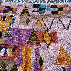May include: A colorful, patterned rug with a light pink background. The rug features geometric shapes in shades of yellow, orange, purple, brown, and black. The rug has a textured appearance and a fringe along the top edge.