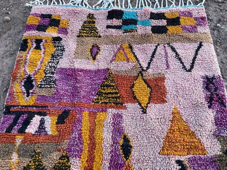 May include: A colorful, patterned rug with a light pink background. The rug features geometric shapes in shades of yellow, orange, purple, brown, and black. The rug has a textured appearance and a fringe along the top edge.