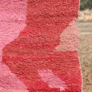 May include: Close-up of a plush, textured rug with a geometric design. The rug features shades of pink and red, with a lighter pink pattern against a darker red background. The rug's texture is visible.