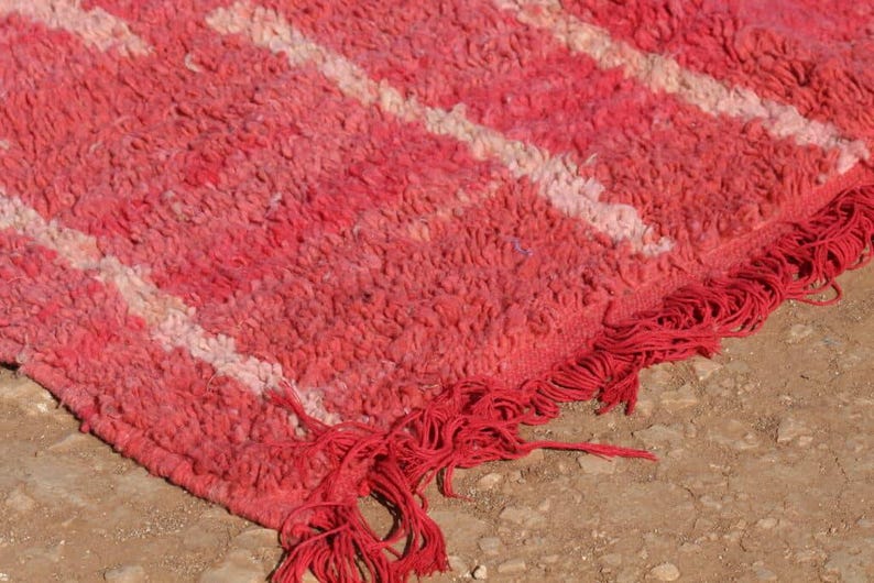 May include: Close-up of a red and white patterned rug with a textured surface. The rug has a fringe along the edge. The rug is made of a thick, woven material. The rug is likely used as a floor covering.