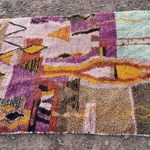May include: A rectangular, multi-colored rug with a geometric design. The rug features a variety of colors, including purple, orange, yellow, and green. The rug has a fringe on one end and is made of a textured material.