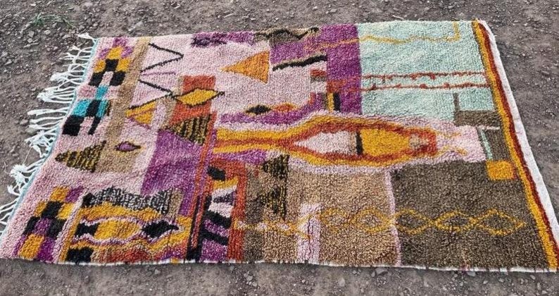 May include: A rectangular, multi-colored rug with a geometric design. The rug features a variety of colors, including purple, orange, yellow, and green. The rug has a fringe on one end and is made of a textured material.
