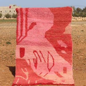 May include: A rectangular, hand-woven rug with a vibrant, abstract design. The rug features a gradient of pink and red hues, with geometric and organic shapes. The rug is displayed outdoors, showcasing its texture and colour variations.