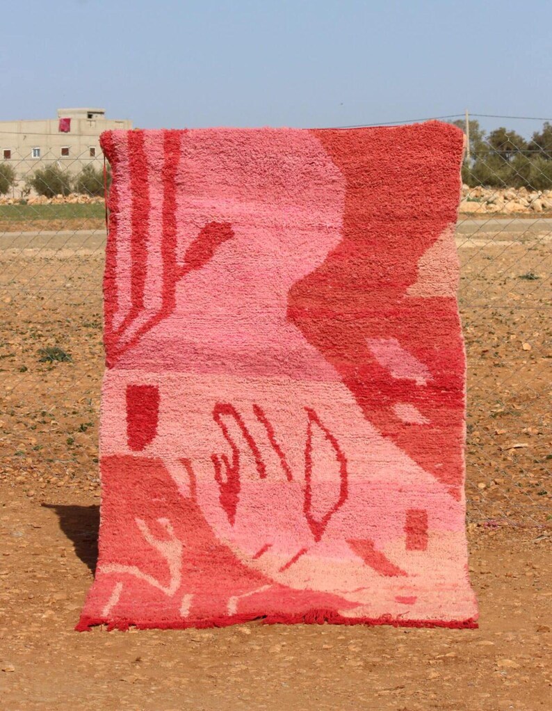 May include: A rectangular, hand-woven rug with a vibrant, abstract design. The rug features a gradient of pink and red hues, with geometric and organic shapes. The rug is displayed outdoors, showcasing its texture and colour variations.