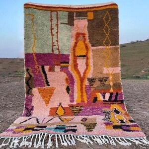 May include: A colorful, abstract rug with a geometric design. The rug features a mix of pink, purple, yellow, and brown hues. It has a fringed edge and is displayed outdoors.