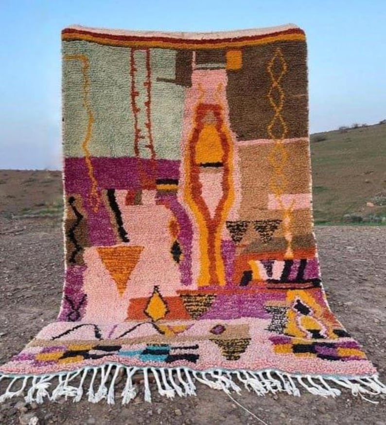 May include: A colorful, abstract rug with a geometric design. The rug features a mix of pink, purple, yellow, and brown hues. It has a fringed edge and is displayed outdoors.