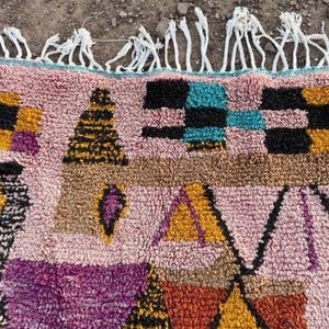 May include: A colorful, patterned rug with a pink background and geometric shapes in black, yellow, purple, and teal. The rug has a textured appearance and white fringe along the top edge. This rug is suitable for home decor.
