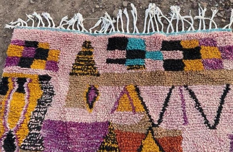 May include: A colorful, patterned rug with a pink background and geometric shapes in black, yellow, purple, and teal. The rug has a textured appearance and white fringe along the top edge. This rug is suitable for home decor.