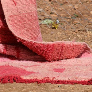 May include: Close-up of a pink and red rug with a textured surface. The rug is folded over, revealing a fringed edge and a pattern of varying shades of pink and red.