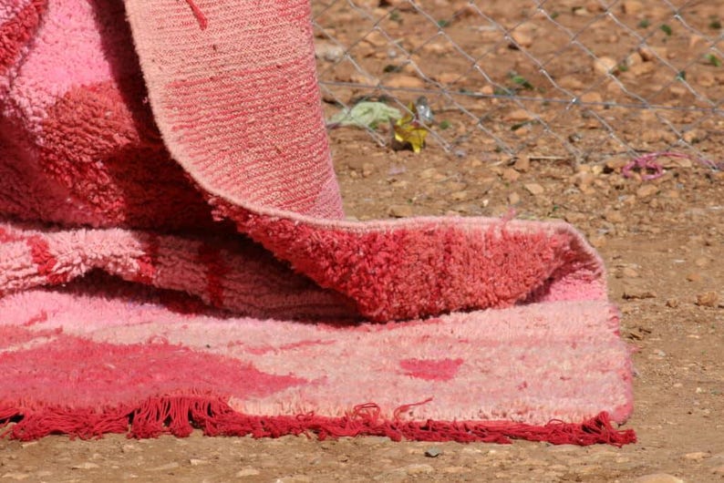 May include: Close-up of a pink and red rug with a textured surface. The rug is folded over, revealing a fringed edge and a pattern of varying shades of pink and red.
