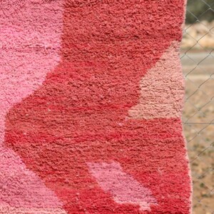May include: Close-up of a plush, textured rug with a geometric design. The rug features shades of pink and red, with a lighter pink pattern against a darker red background. The rug's texture is visible.