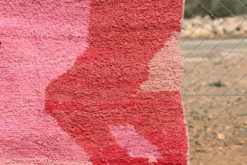 May include: Close-up of a plush, textured rug with a geometric design. The rug features shades of pink and red, with a lighter pink pattern against a darker red background. The rug's texture is visible.