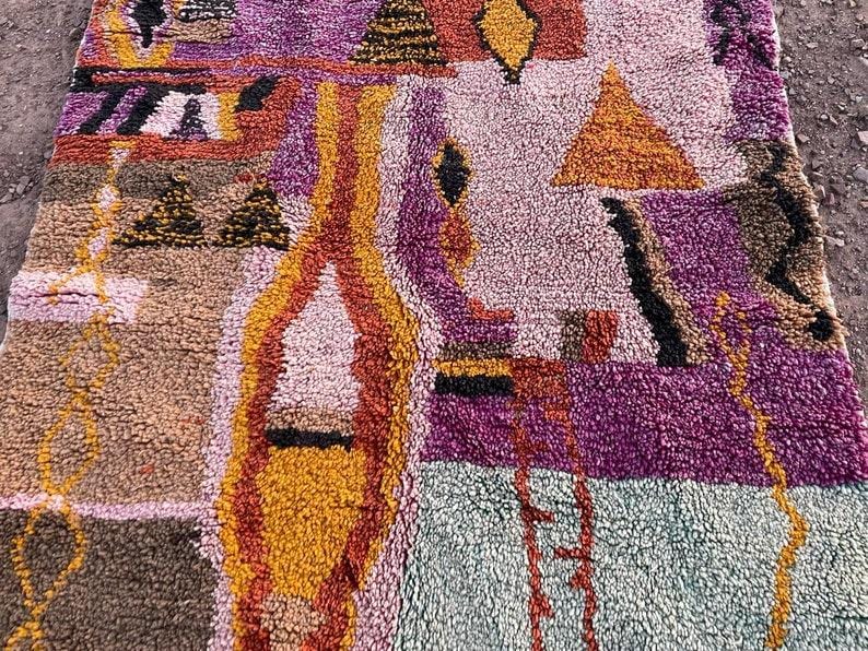 May include: A colorful, textured rug with a geometric design. The rug features a mix of colors, including purple, orange, yellow, and brown. The design includes triangles, diamonds, and other shapes. The rug is made of a thick, woven material.
