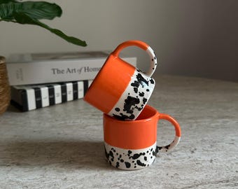 May include: Two orange ceramic mugs stacked on top of each other. The top half of each mug is orange, while the bottom half features a white base with black speckled detailing. The handles are white with black speckles.