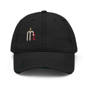 May include: A black distressed baseball cap featuring a small graphic on the front panel. The design consists of a white letter M with a gold wheat stalk icon above it and a small red curved shape attached to the right side of the letter.