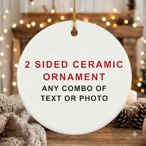 May include: A white, round ceramic ornament with gold hanging string. The ornament has the text "2 SIDED CERAMIC ORNAMENT ANY COMBO OF TEXT OR PHOTO" in red and black. The background includes a Christmas tree and lights.