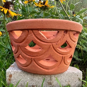 May include: A terracotta planter with a wide, circular rim and a repeating scalloped cutout pattern across its exterior surface. The clay pot features a matte, earthy orange finish and a hollow interior, designed with multiple open arches.