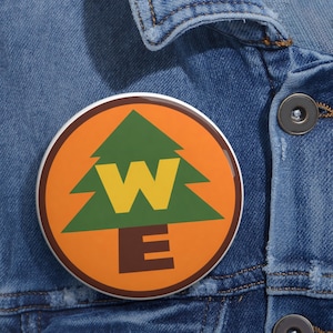 May include: A circular pinback button featuring an orange background with a dark brown circular border. Centered is a green pine tree graphic with a large yellow letter W overlaid on the tree and a brown letter E below the tree.