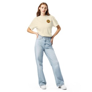 May include: A cream-coloured short-sleeve t-shirt featuring a small circular patch on the left chest. The patch displays a green pine tree graphic with a yellow letter W inside an orange border. The shirt is paired with light blue denim jeans.